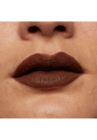 Matte brun läppstift applicerat på något fylliga läppar. Texturen visar fina linjer, med en slät finish och jämn färgfördelning.