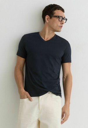 Mand iført sort V-hals T-shirt og hvide bukser, står mod en hvid væg og kigger til højre med den ene hånd i lommen.