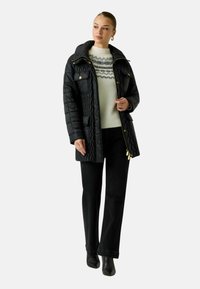 Giacca imbottita nera trapuntata con accessori dorati, tasche frontali e cappuccio. Indossata sopra un maglione crema con motivo geometrico e pantaloni neri.