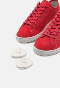 Zapatillas de ante rojo con suelas y ojales de goma blanca, con cordones rojos a juego y dos pares adicionales de cordones blancos.