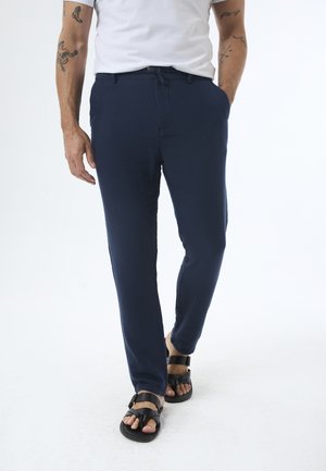 Pantaloni blu navy in un tessuto leggero, caratterizzati da una vestibilità slim, tasche laterali e vita con cordino. Abbinati a sandali neri con laccetti.