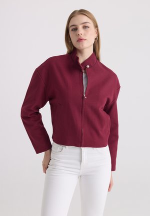 Femme portant une veste bordeaux à fermeture éclair et col montant ainsi qu'un pantalon blanc, debout devant un fond clair uni avec une main derrière le dos.