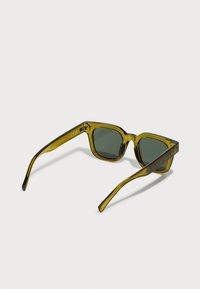 Gafas de sol con tintado verde y un armazón de acetato grueso y transparente en color verde oliva. Lentes cuadradas, varillas rectas y detalles de herrajes mínimos.