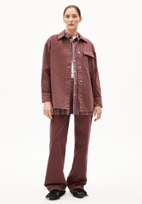 Frau in lockerem burgunderrotem Jacket und Hose über gestreiftem Shirt und weißem Top, stehend vor schlichtem weißen Hintergrund.