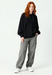 Felpa nera con cappuccio, tasca frontale e lacci, abbinata a pantaloni cargo grigi larghi con tasche laterali. Sneakers bianche completano l'outfit.