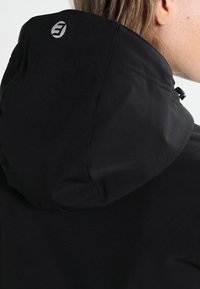 Veste noire à capuche avec une texture lisse, un cordon de serrage ajustable et un petit logo au dos de la capuche.