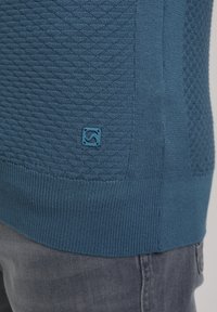 Pull en maille texturée bleu avec un petit patch logo carré près de l'ourlet, porté avec un jean en denim gris.