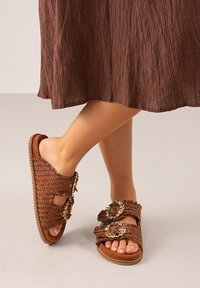 Sandalias tejidas marrones con correas texturizadas, adornadas con cuentas de colores, que presentan un diseño de punta abierta y una suela plana de goma.