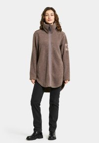 Brun fleece oversized jakke med høj krave, fuld frontlynlås og buet kant. Har et logo-patch på venstre arm. Bæres med sorte bukser.