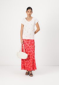 Witte katoenen t-shirt met kleine, kleurrijke prints, gecombineerd met een rode rok met bloemenpatroon. Het model houdt een witte handtas vast en draagt zwarte sandalen.