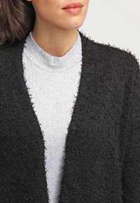 Cardigan nero con texture a rilievo, superficie pelosa, con apertura frontale; indossato sopra un dolcevita grigio chiaro a costine. Design semplice senza chiusure.