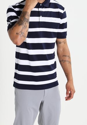 Mand iført en marineblå og hvid stribet poloshirt og lysegrå bukser, som viser tatoveringer på begge arme.