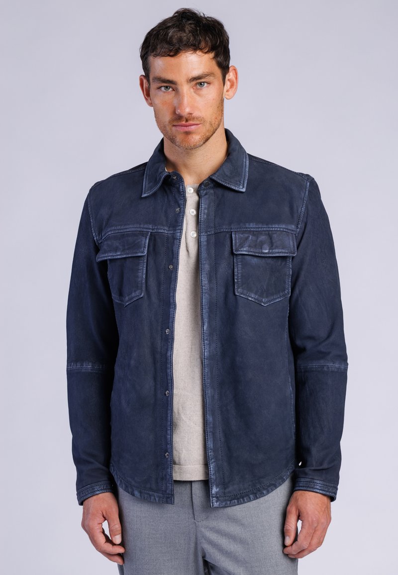 Hombre con chaqueta abotonada azul oscuro sobre una camisa beige clara y pantalones grises, mirando hacia adelante sobre un fondo liso.