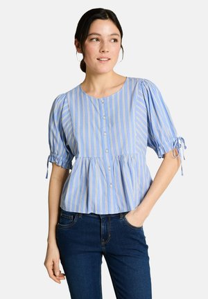 Frau trägt eine blau-weiß vertikal gestreifte Bluse mit kurzen Puffärmeln und dunkelblauen Jeans, Hand in der Hosentasche.