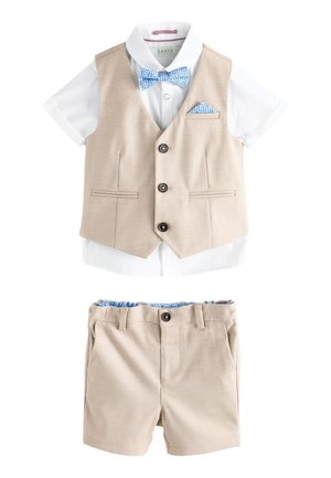 Mladý chlapec nosi béžovú vestu a krátke nohavice, bielu krátky rukáv košeľu, modrý vzorovaný motýlik a vreckovku, stojí na sivom podlahe.