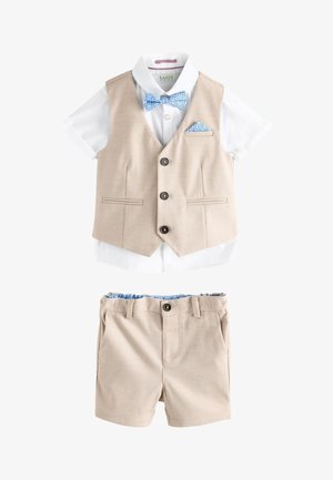 Jongentje met beige vest en korte broek, wit overhemd met korte mouwen, blauw patroon vlinderdasje en pochet, staand op een grijze vloer.