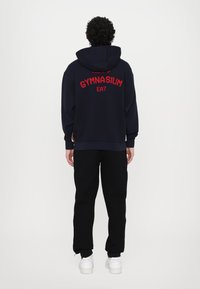 Marinblå hoodie med röd text "GYMNASIUM EA7", elastiska mudd och nederkant; ihop med svarta sweatpants och vita sneakers. Enfärgad bakgrund.