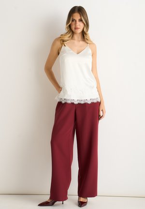 Woman wearing white lace-trim camisole and wide-leg burgundy pants, standing with hand in pocket against plain background.