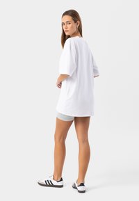 Biela oversize tričko s krátkymi rukávmi, vyrobené z bavlny. Má okrúhly výstrih a je kombinované so šedými šortkami a čierno-bielymi topánkami.