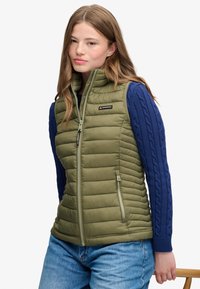 Jeune femme aux longs cheveux portant une veste matelassée vert olive sur un pull bleu à torsades et un jean bleu clair, debout avec une main posée sur une chaise.