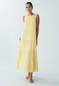 Geltona maxi suknelė su apvalia iškirpte, be rankovių ir sluoksniuotais raukiniais apačioje. Lygi medžiaga, laisvo kirpimo ir kulkšnies ilgio.