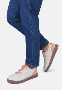 Baskets en toile blanches avec des accents orange et rose, semelle en caoutchouc texturée, bout rond et lacets violets, assorties à un pantalon en denim bleu.