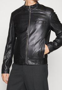 Veste en cuir noir avec un col montant, des coutures verticales sur les épaules, deux poches zippées, et un design élégant et ajusté.