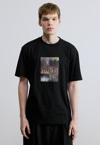 Μαύρο βαμβακερό T-shirt με ορθογώνιο γραφικό τύπωμα που αναγράφει το "HUGO" και έχει υφασμάτινο φόντο σε γήινους τόνους. Κοντά μανίκια.