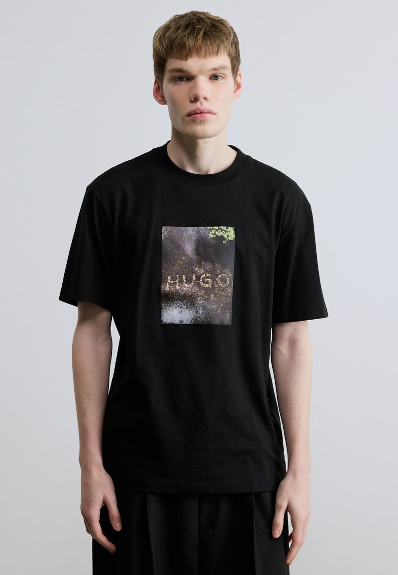 Μαύρο βαμβακερό T-shirt με ορθογώνιο γραφικό τύπωμα που αναγράφει το "HUGO" και έχει υφασμάτινο φόντο σε γήινους τόνους. Κοντά μανίκια.