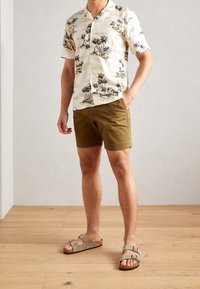 Korte mouw beige shirt met palmboomprint, gecombineerd met olijfgroen shorts en beige strap sandalen op een houten vloer achtergrond.
