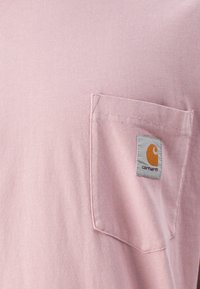 Svetlo roza majica s kratkimi rokavi z mehko teksturo in žepom na levi strani prsnega dela, ki ima oznako Carhartt v oranžni in sivi barvi.