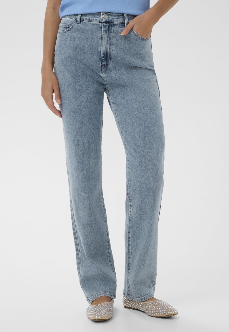 Woman wearing light blue straight-leg jeans with hand in pocket and beige studded ballet flats, standing against a plain background.