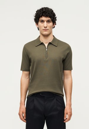 Ung mand med krøllet hår iført en olivengrøn kortærmet polo med lynlås foran og sorte bukser, der står mod en ensfarvet baggrund.