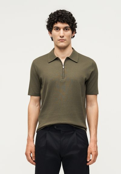 Jeune homme aux cheveux bouclés portant un polo vert olive à manches courtes avec une fermeture éclair à l'avant et un pantalon noir, debout devant un fond uni.