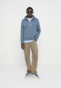 Μπλε φούτερ με φερμουάρ στον γιακά πάνω από ένα ανοιχτό μπλε ριγέ πουκάμισο, συνδυασμένο με μπεζ παντελόνι και γκρι αθλητικά παπούτσια. Φοράει γυαλιά ηλίου.