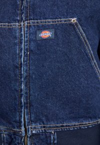 Jeansjakke i mørkeblå, med lynlås, ribbede manchetter og en brystlomme med et Dickies-logo patch. Distinktiv syning.