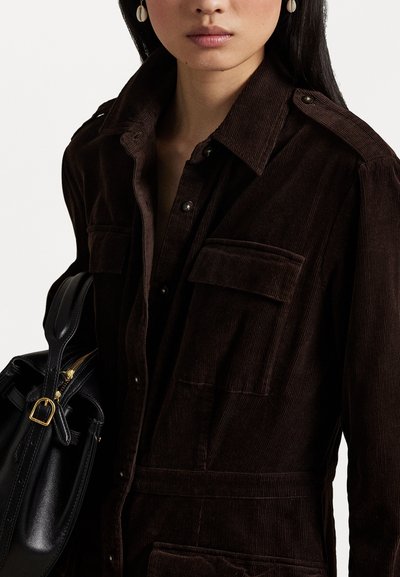 Bruine corduroy jurk met schouderepauletten, twee borstzakken en een ceintuur in de taille. Gepaard met een zwarte handtas met gouden accenten.