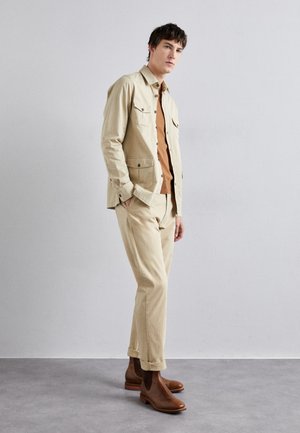 Veste en coton beige avec deux poches poitrine, portée sur une chemise marron et un pantalon beige assorti. Des bottines en cuir marron à la cheville viennent compléter la tenue.