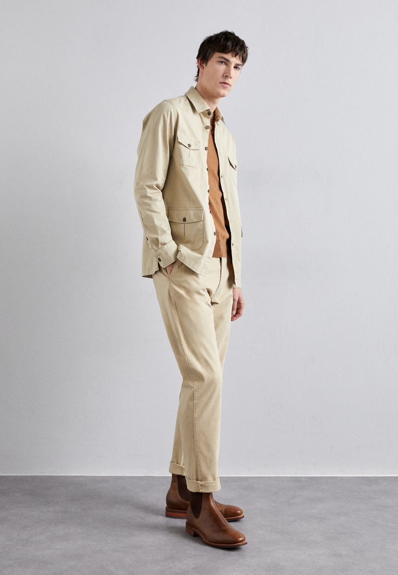 Chaqueta de algodón beige con dos bolsillos en el pecho, sobre una camisa marrón y pantalones beige a juego. Botas de cuero marrón hasta el tobillo completan el atuendo.