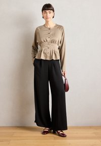 Beige satinbluse med frontknapper og samlet talje, satt sammen med svarte vide bukser. Modellen holder en liten rød veske og har på seg burgunder hæler.
