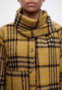 Personne portant un manteau en laine à carreaux jaune, noir et beige avec un col haut plissé et de grands boutons.
