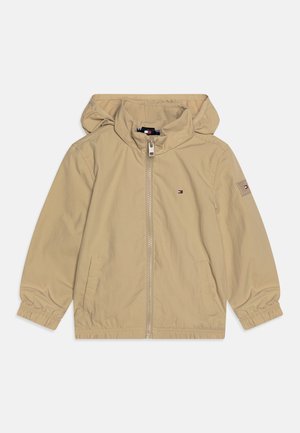 Giacca leggera beige con cappuccio, polsini elasticizzati, zip frontale, due tasche laterali e un piccolo logo sul lato sinistro del petto. Tessuto liscio.