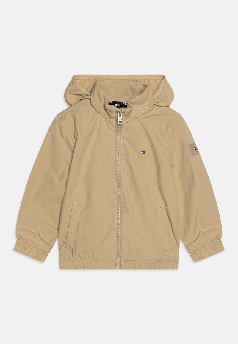 Beige lätt jacka med huva, elastiska manschester, frontdragkedja, två sidofickor och en liten logotyp på vänster bröst. Slät textur.