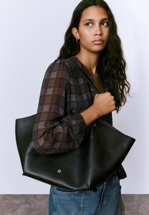 Black leather tote bag with a textured finish, wide opening, and a subtle logo. Model wears a checkered blouse and denim jeans.