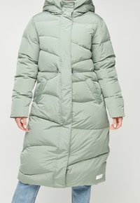 Vihreä puhvitulistin, jossa on huppu, diagonaaliseen ompeleeseen, kaksi sivutaskua ja etuvetoketju. Käytetään farkkujen päällä, keskipitkä malli.