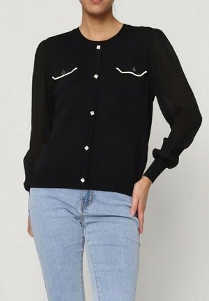 Blouse noire à manches longues en voile, avec des détails blancs sur les poches et des boutons métalliques carrés. Portée avec un jean bleu clair.