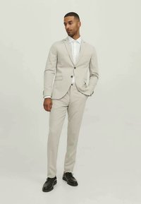 Fato cinza claro de duas peças com um design ajustado, fecho de um botão, lapelas tipo notch e uma camisa branca por baixo. Sapatos pretos completam o visual.