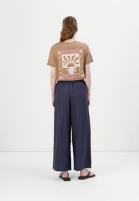 Bruin T-shirt met grafische print "Sunset Seeker" op de achterkant, gecombineerd met losse marineblauwe wijde broek en zwarte platte sandalen.