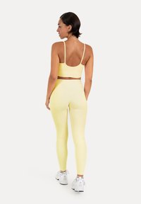 Femme aux cheveux courts et foncés, portant un soutien-gorge de sport jaune et des leggings, baskets blanches, debout, tournée de dos sur un fond blanc.