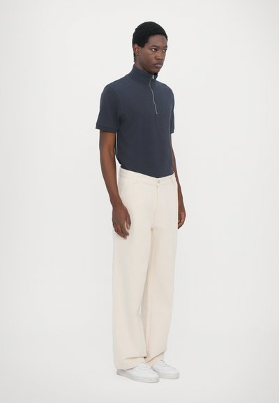 Navy zip-up polo shirt with short sleeves paired with cream wide-leg trousers and white sneakers. Plain background highlights the outfit.